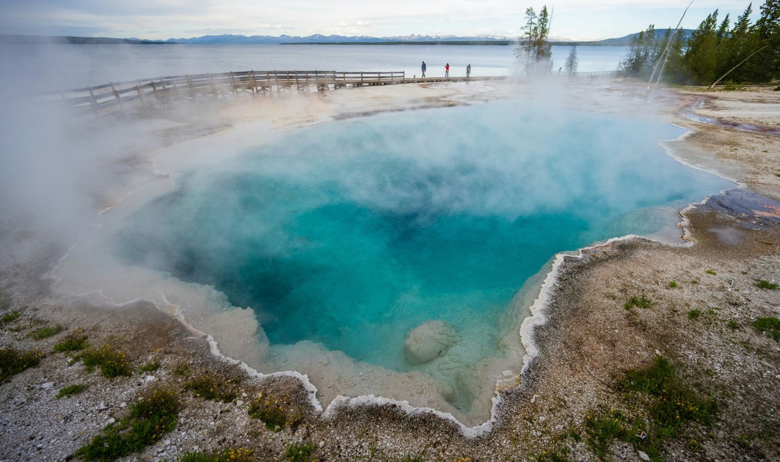 A wide turquoise hot spring pool with steam drifting across it, its rim crusted pale and the ground round it bare and stony with small yellow flowers, a wooden boardwalk along the far side with three people standing on it, a lake beyond, and a line of mountains under a grey sky.