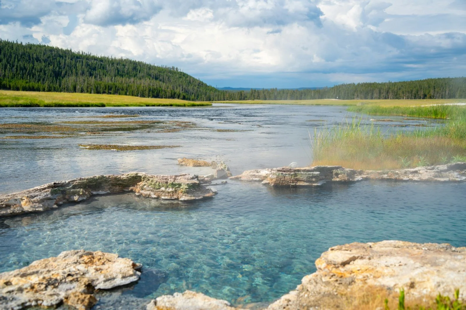 A clear turquoise hot spring pool held in a rim of pale mineral rock, steam drifting off it, beside a wide shallow river with grass banks and a dark pine-covered hill under a cloudy sky.