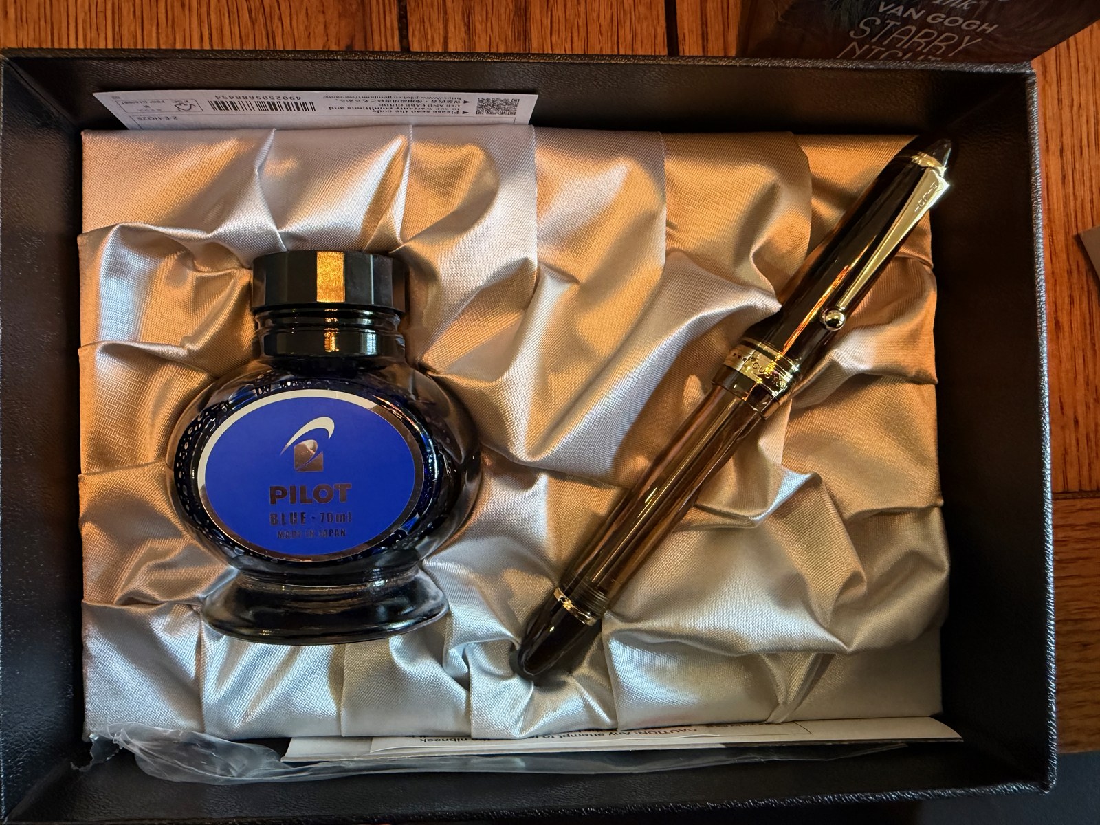An open black presentation box lined with folded silver satin, holding a round blue glass bottle of ink whose label reads PILOT, BLUE, 70ml, and a capped dark brown fountain pen with gold trim.