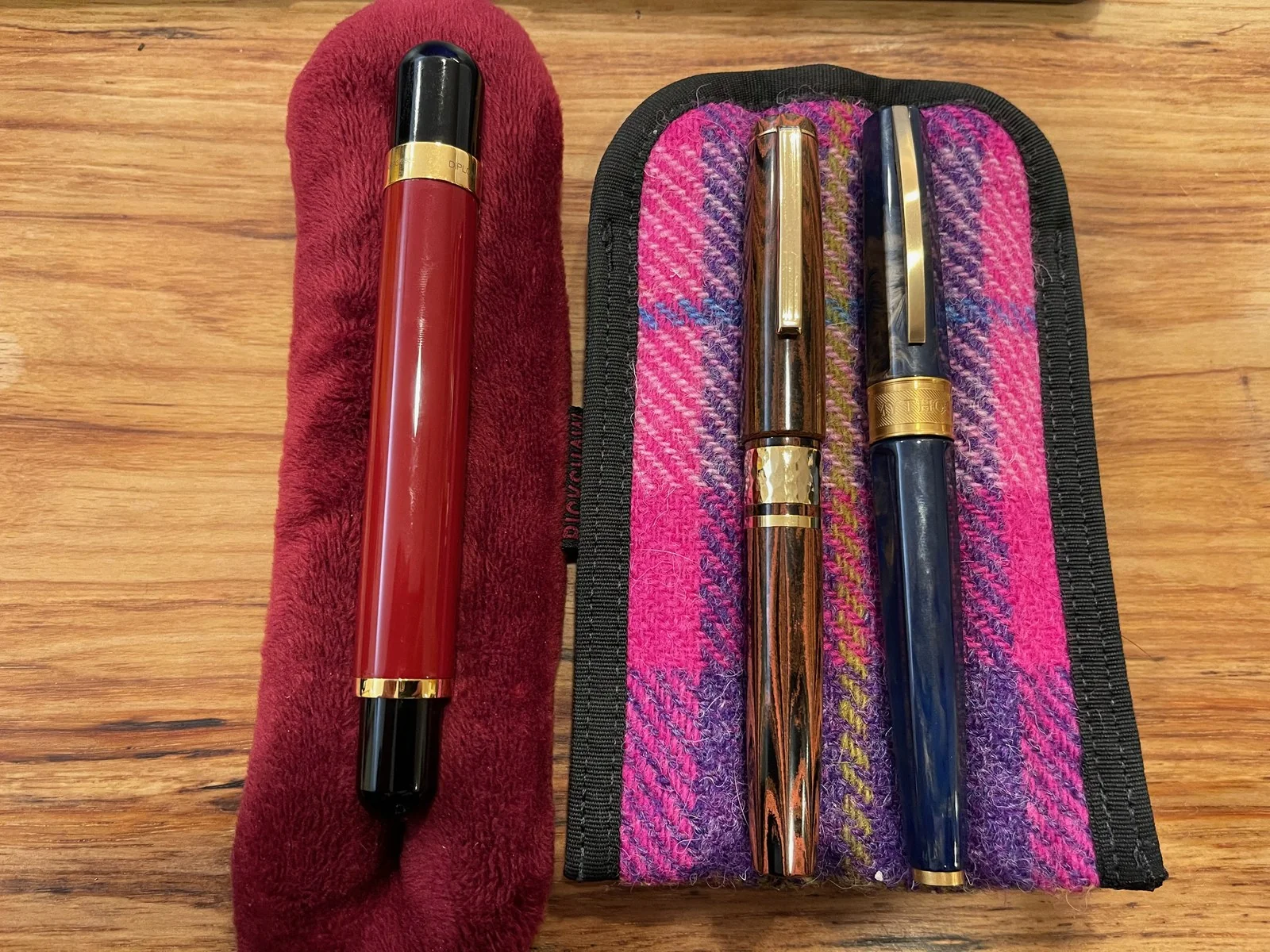 On a wooden table, a red and black fountain pen with gold bands lies on a plush burgundy sleeve, beside a pink and purple tweed case holding a copper-and-black striped pen and a marbled navy blue pen, both with gold clips.
