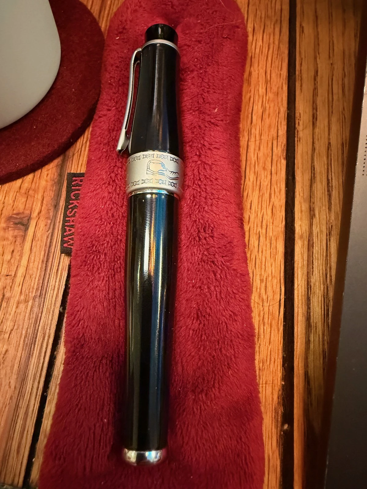 A glossy black fountain pen with a silver clip lies on a red plush sleeve whose tab reads RICKSHAW. Its satin silver band carries a small engraved drawing between two rows of tiny squares.