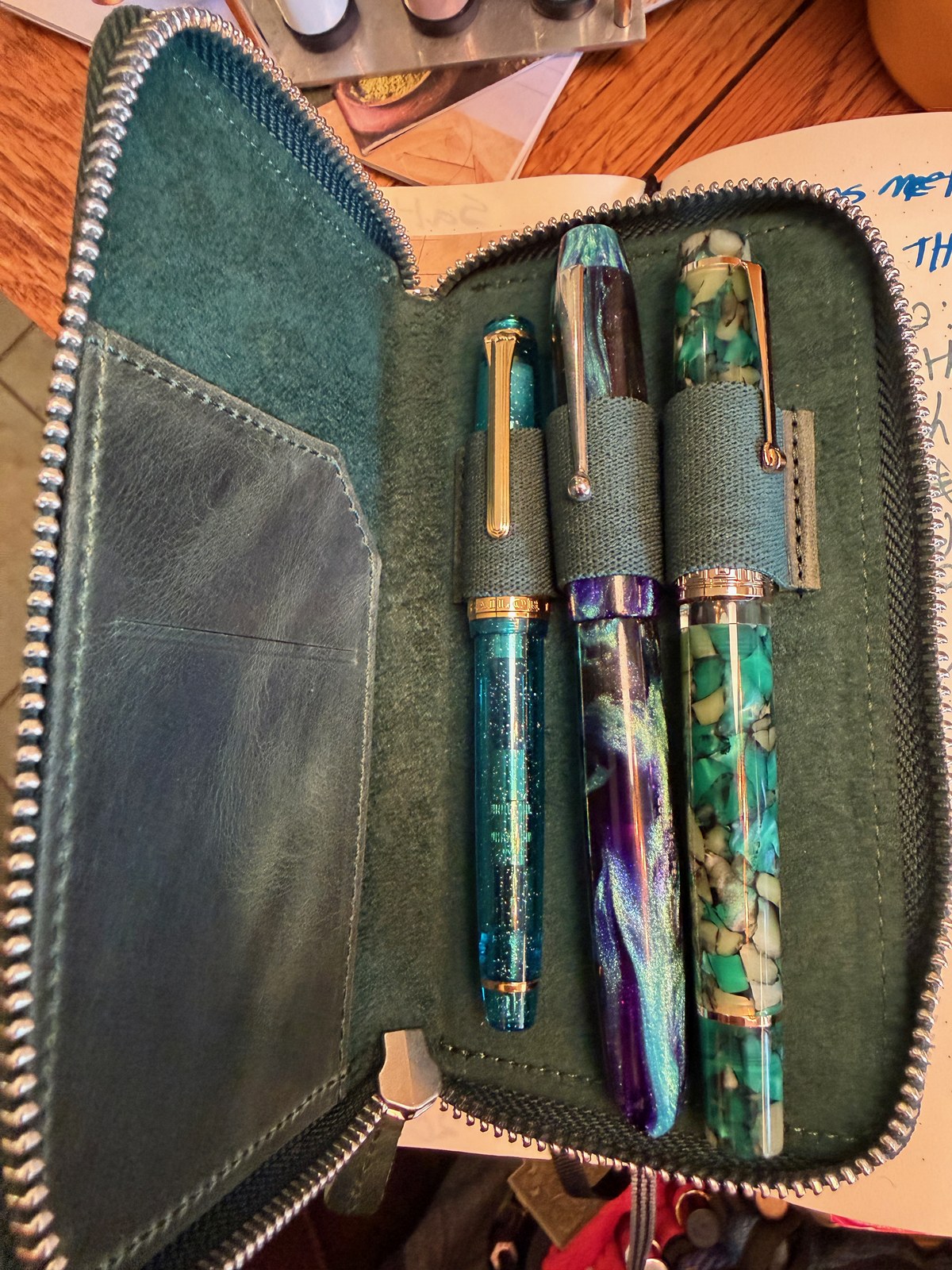 A dark green leather case lies unzipped, with pens held in its elastic loops: a clear turquoise pen full of sparkle with a band reading SAILOR, a purple and teal swirl that shimmers, and a green and cream pen like broken stone.