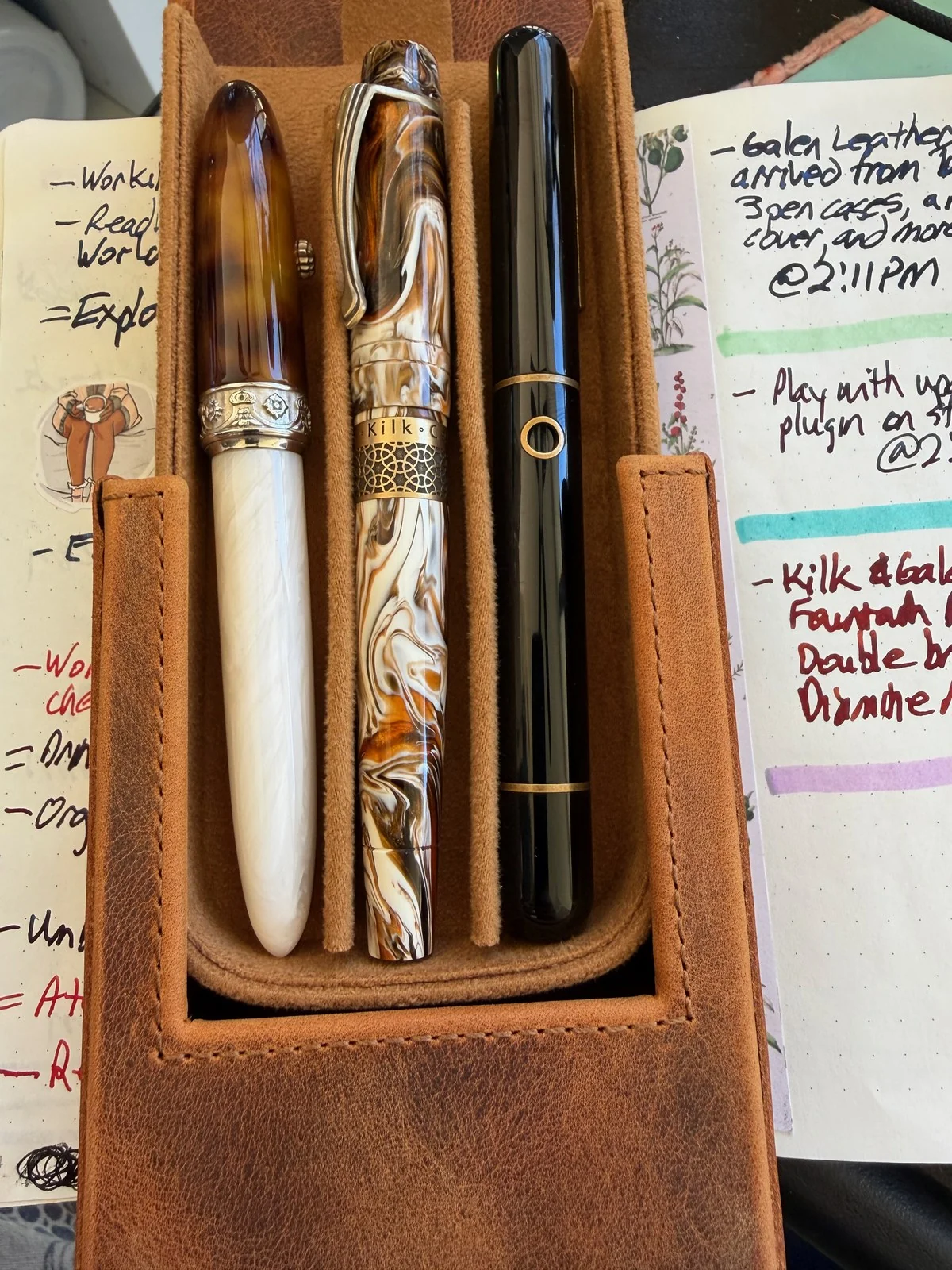 A tan leather pen case, flap open over a handwritten journal. In it: a tortoiseshell cap on a pearly white body with an ornate silver band, a swirled brown, cream and orange pen whose band reads Kilk, and a glossy black pen with gold rings.