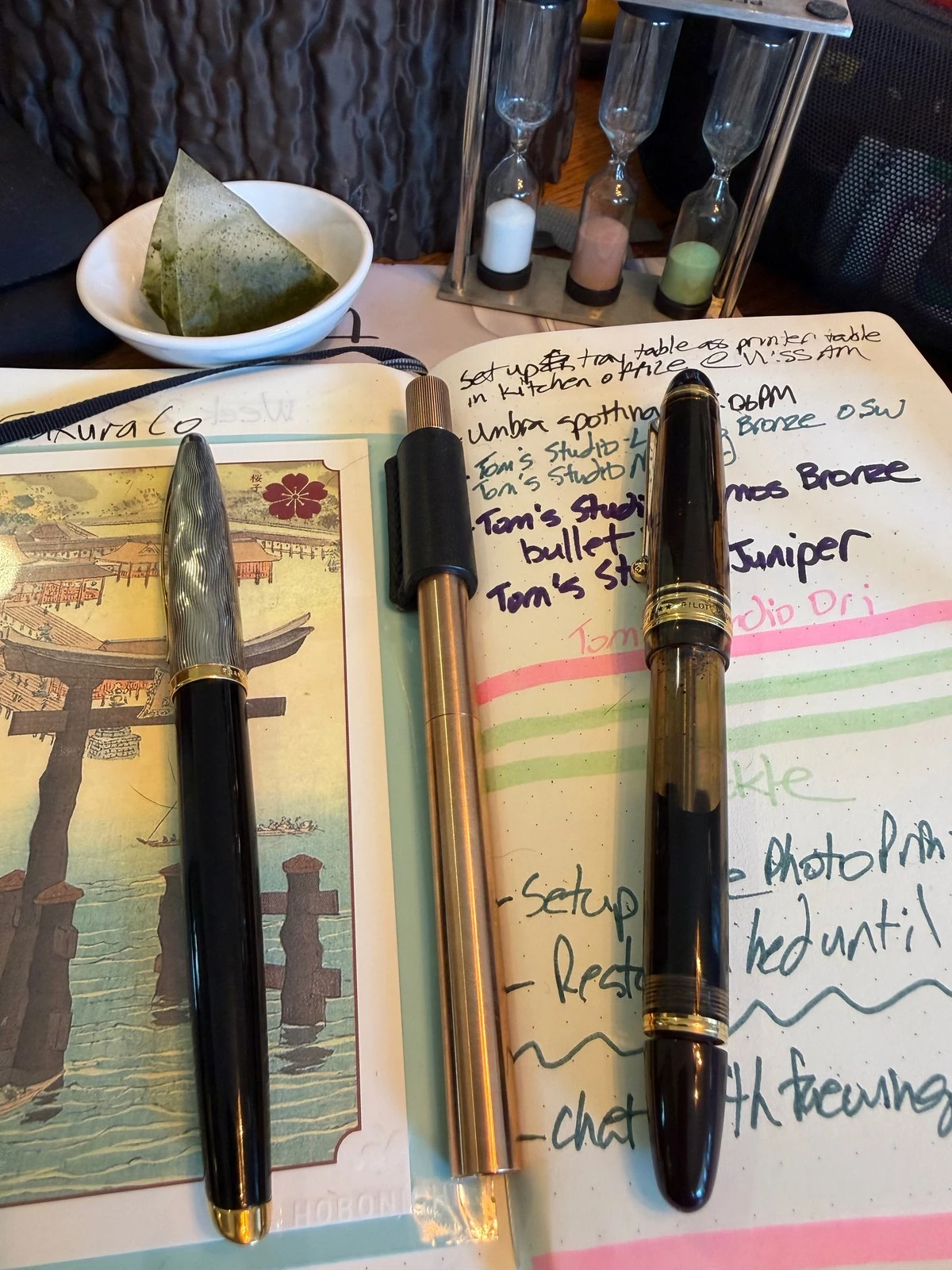 An open journal with a print of a red torii gate in water on one page and notes in coloured inks on the other. On it: a black pen with a wavy silver cap, a slim brass pen, and an amber see-through pen reading PILOT.