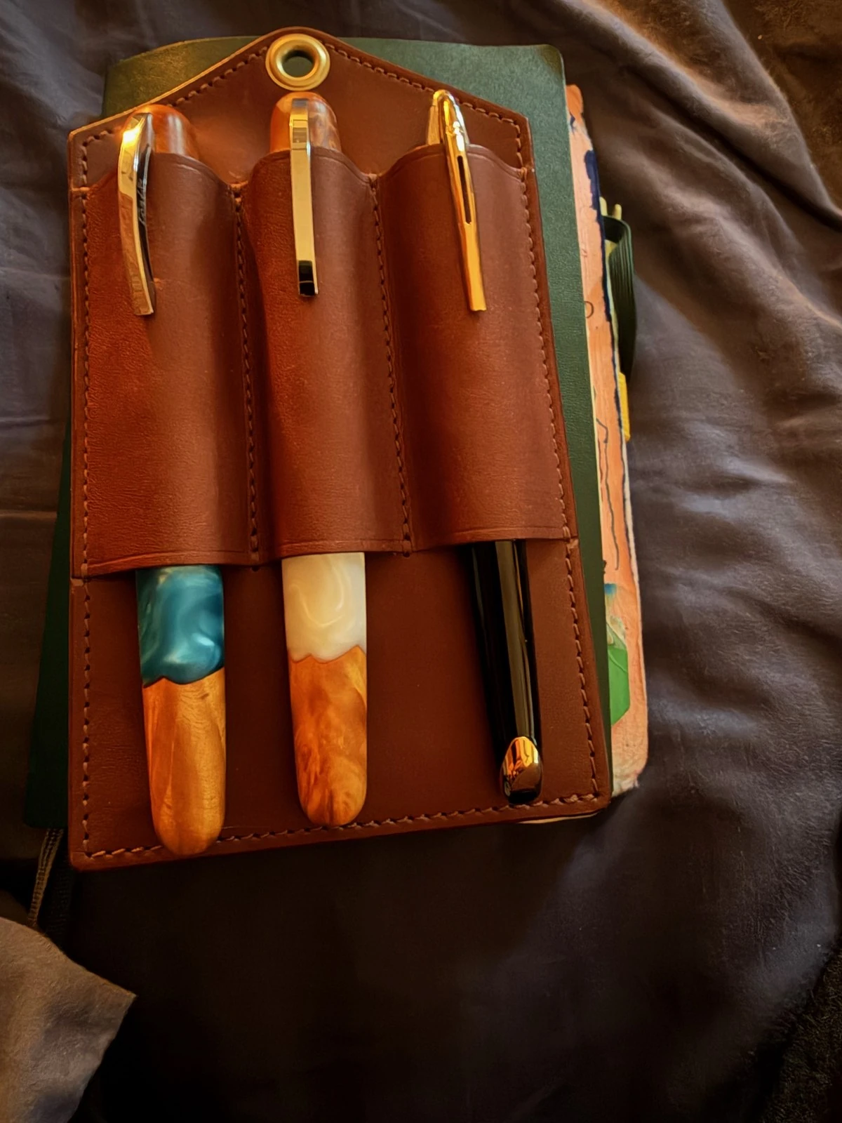 A brown leather holder with stitched slots sits on a green notebook on a rumpled grey blanket. Two of its pens are cast half in turquoise or pearly white and half in glowing orange-gold; the third is black with a gold clip.