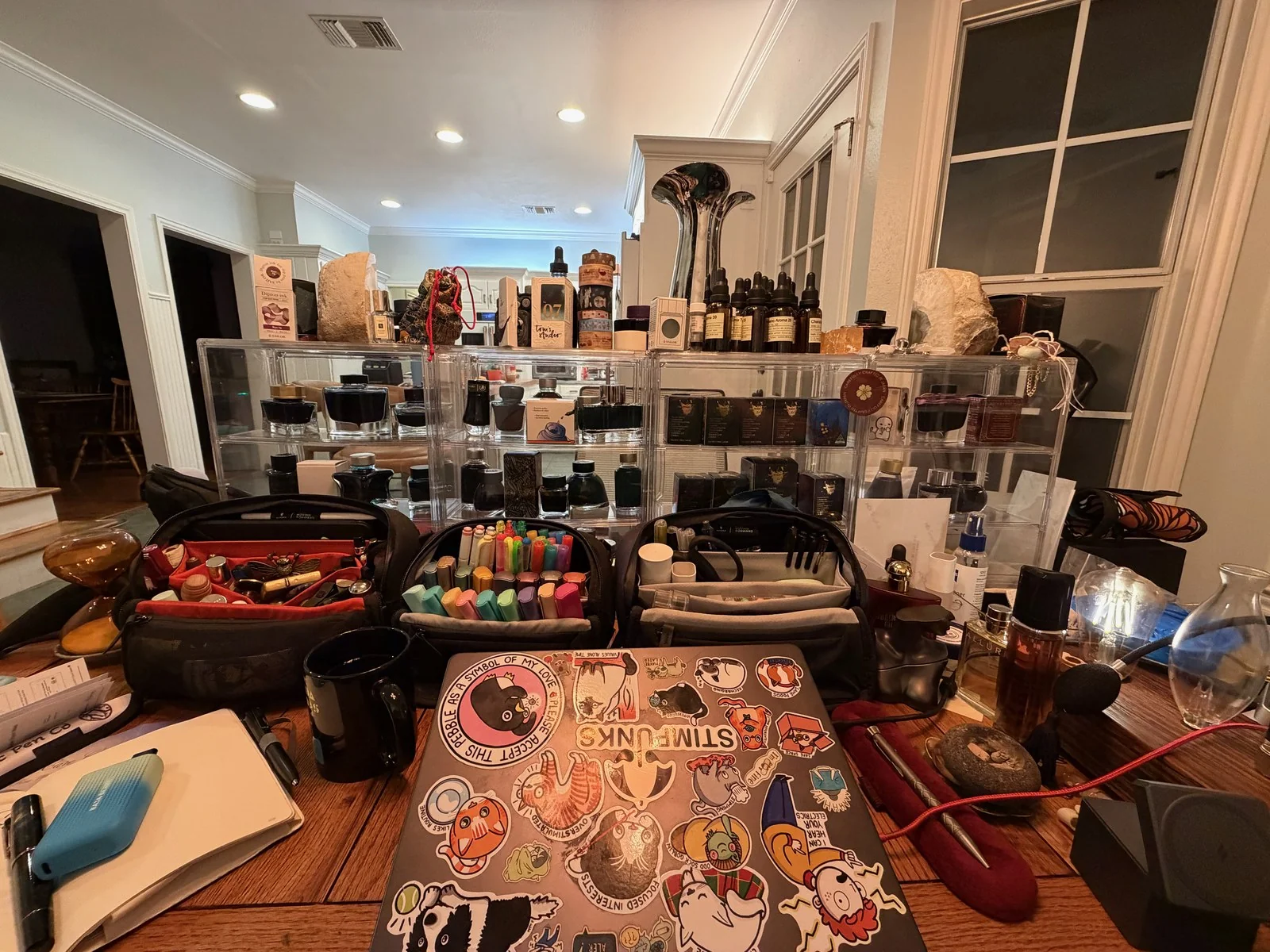 A wide view across a wooden table at night: clear acrylic shelves stacked with ink bottles and boxes, open bags packed with pens and markers, perfume bottles, rocks and sand timers, and in front a laptop covered in stickers, one reading STIMPUNKS.