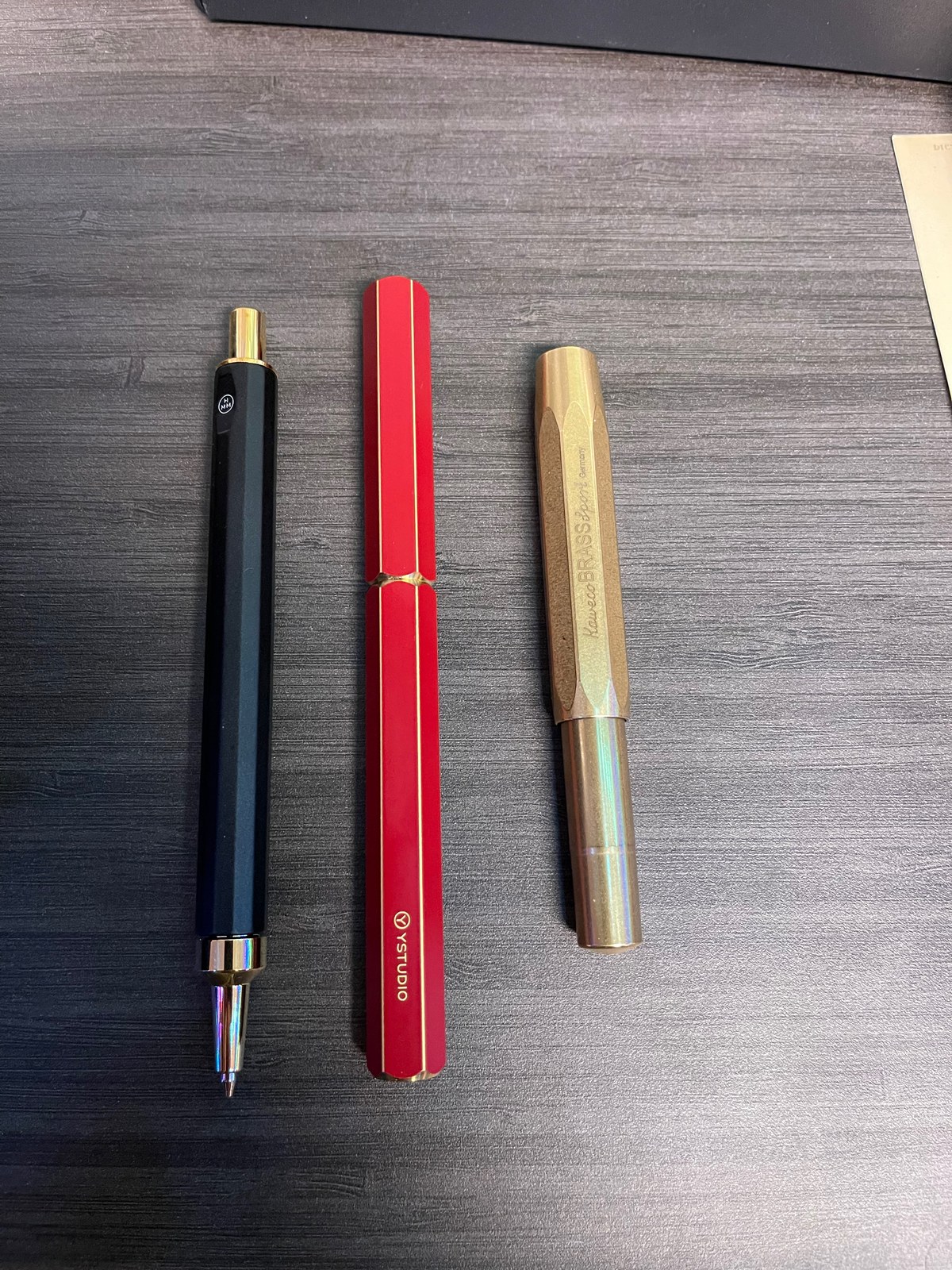 Pens side by side on a dark grey wood-grain desk: a black hexagonal pen with gold ends and its tip out, a red faceted pen with thin gold pinstripes that reads YSTUDIO, and a faceted brass pen that reads Kaweco BRASS Sport Germany.