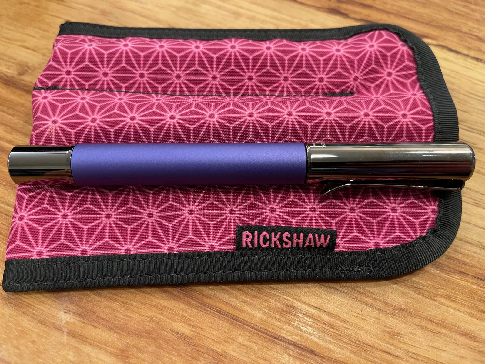 A matte purple pen with a glossy gunmetal cap and clip lies across a pink fabric pen sleeve printed with a lattice of six-pointed stars, with a black tab that reads RICKSHAW.