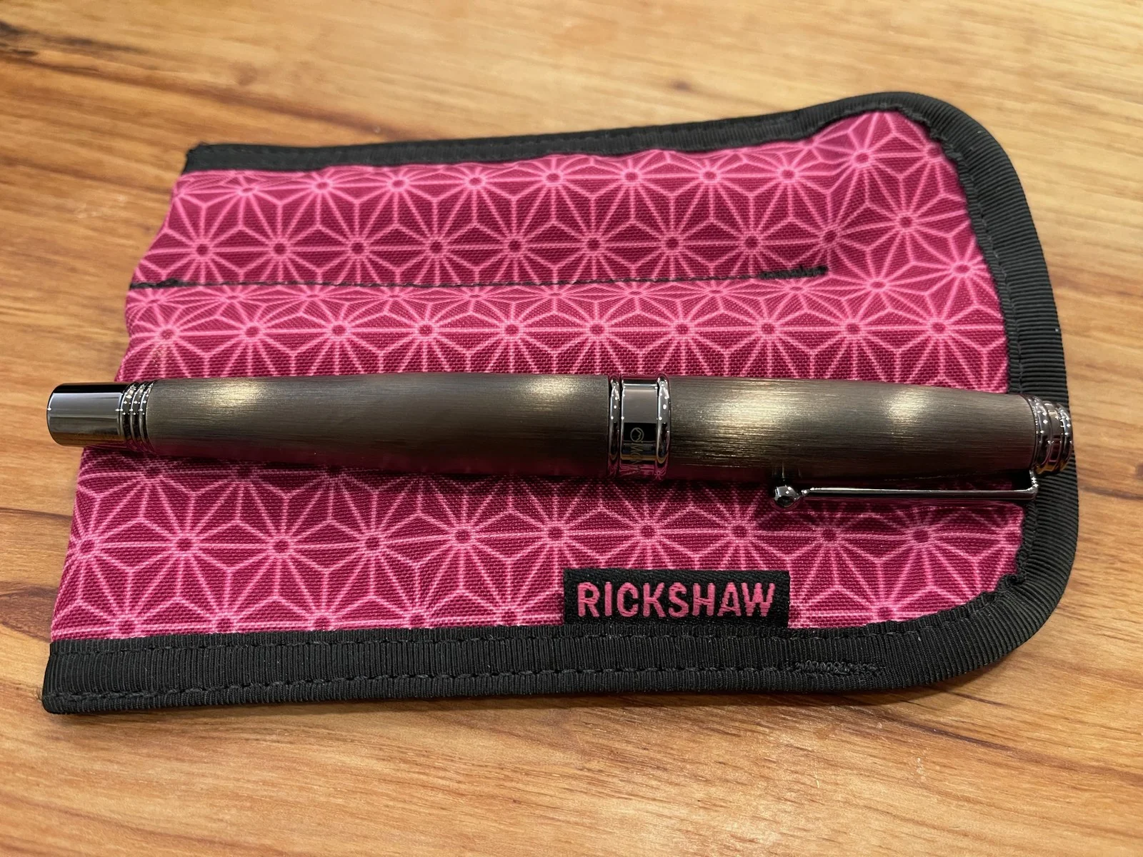 A brushed gunmetal pen with dark chrome bands, clip and ends lies across a pink fabric pen sleeve printed with six-pointed stars, its black tab reading RICKSHAW.