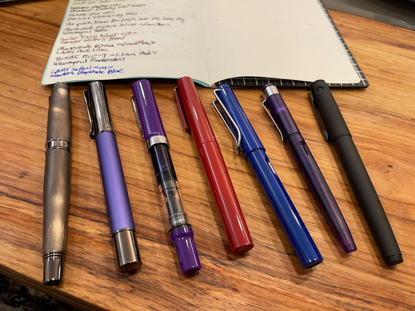 Fountain pens in a row on a wooden desk, under an open notebook listing pens and inks in handwriting, each line in its own colour: brushed gunmetal, matte purple, a purple see-through demonstrator, glossy red, bright blue, translucent purple and matte black.