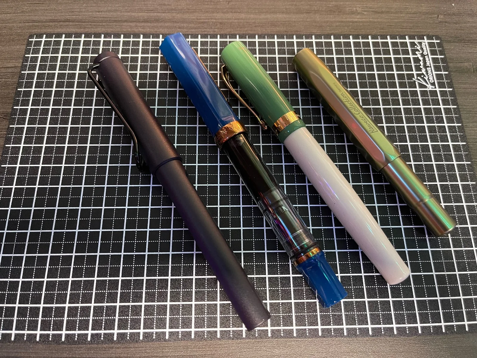 Pens on a black cutting mat ruled in white: a matte navy pen with a wire clip, a blue and clear see-through pen with rose-gold trim, a sage green pen with a pearly white body, and a faceted metal pen shifting green to bronze.