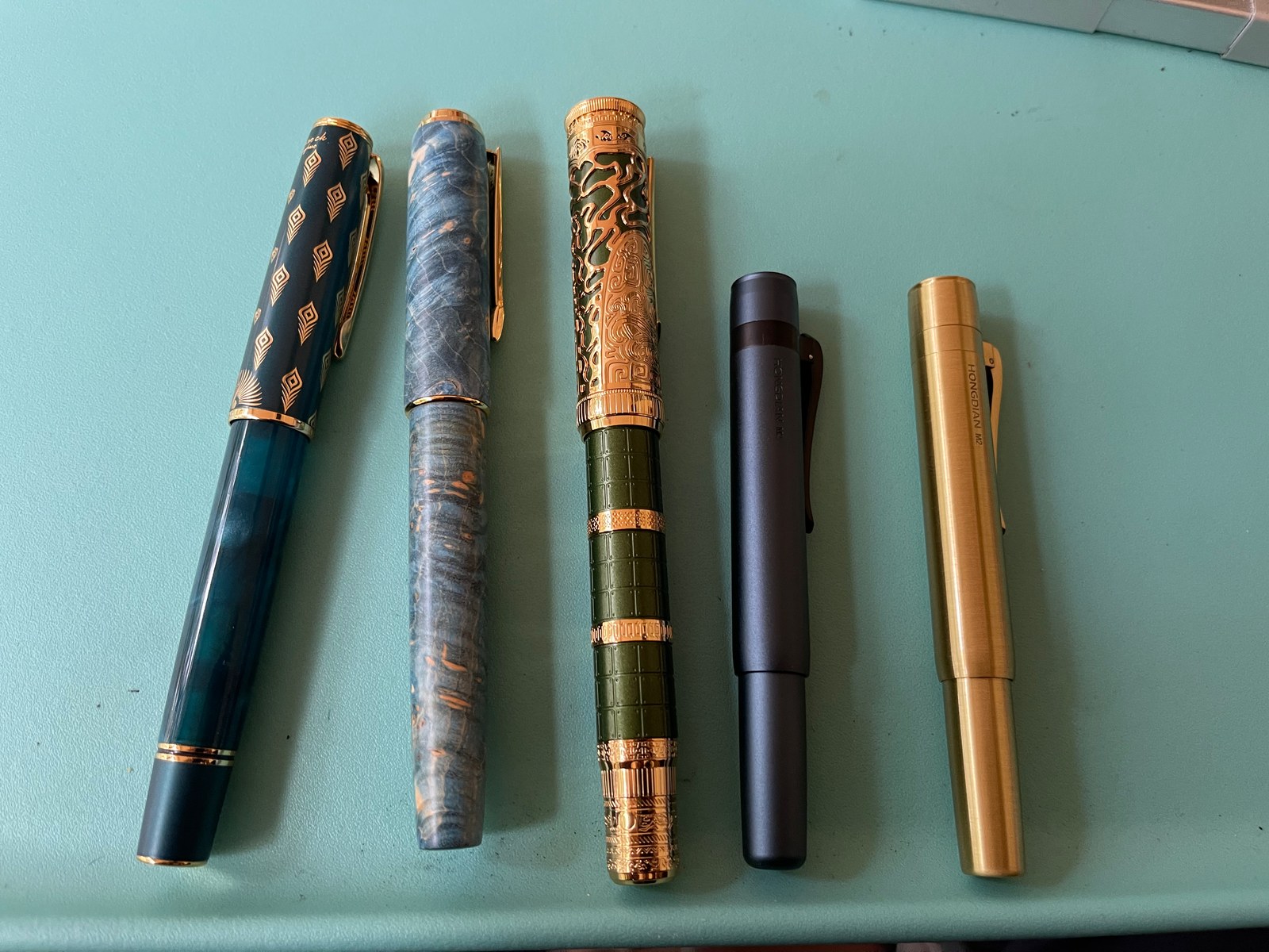 Pens on a turquoise tabletop: a teal pen with gold peacock feathers on its cap, one patterned like blue and gold burl, an ornate green pen with an openwork gold cap, a slim dark grey metal pen, and a brass pen reading HONGDIAN M2.