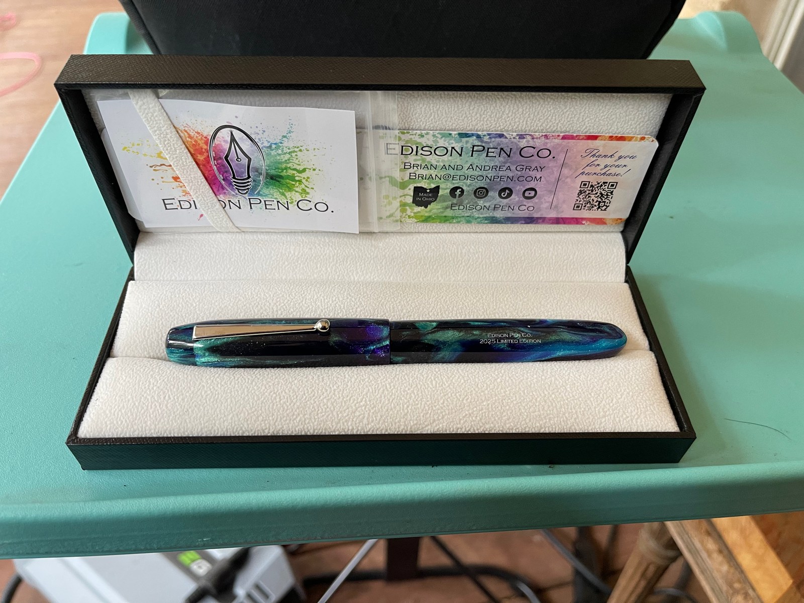 An open black pen box lined in white holds a shimmering fountain pen swirled in deep purple, blue and teal, with a silver clip and the words Edison Pen Co. engraved on its barrel. Behind it, the maker's cards are splashed with rainbow paint.