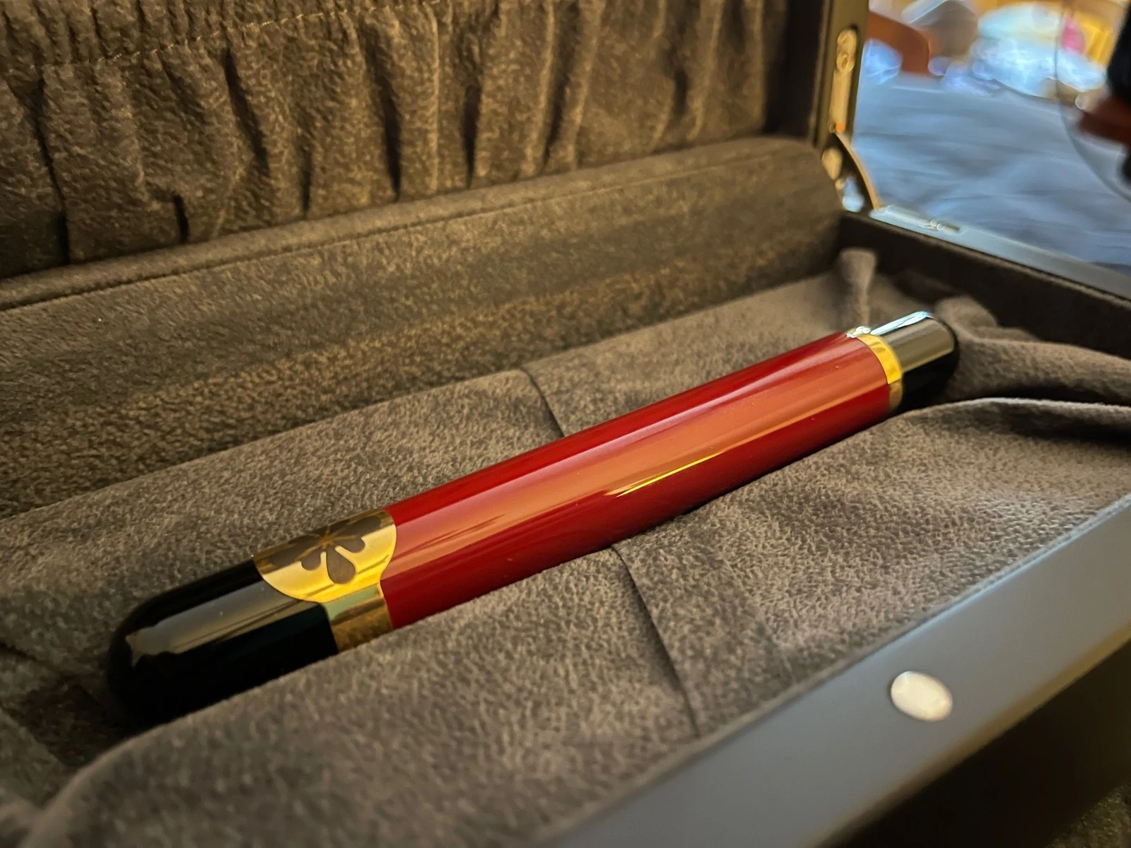 A close view of a glossy red and black fountain pen resting in a box lined with grey suede, with a gold band near one end cut with a black flower.