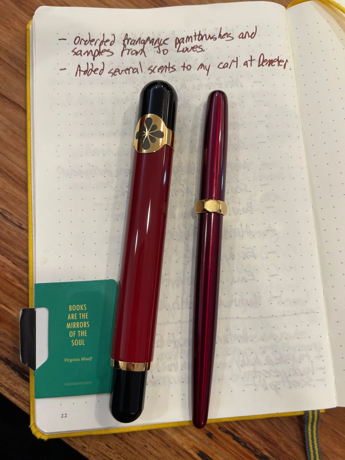 On an open dotted journal lie a red and black pen with a gold band set with a black flower and a slim glossy dark red pen with a faceted gold ring. A green bookmark quotes Virginia Woolf: books are the mirrors of the soul.