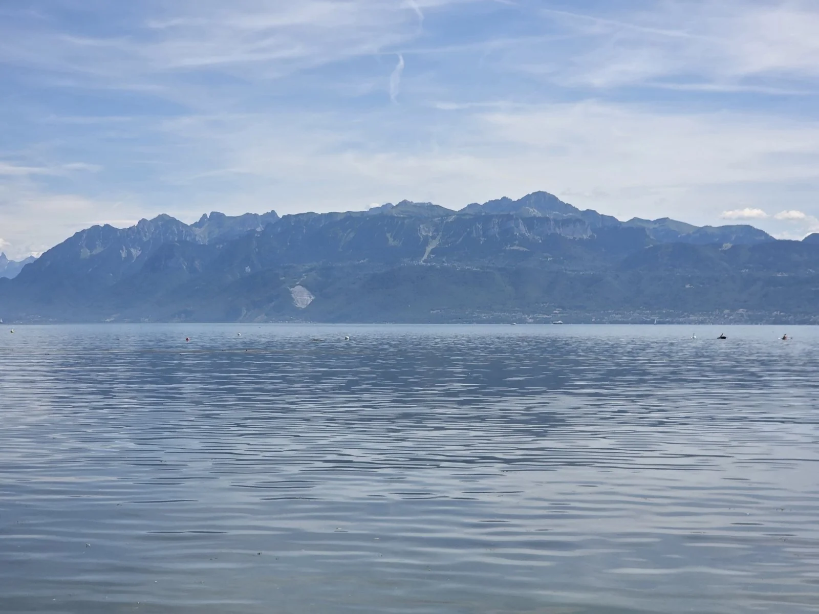Open lake water, rippled and blue-grey, with a row of small buoys in the distance, the far shore’s mountains rising steep and green-grey with a pale scar on one slope, and thin white contrails across a blue sky.