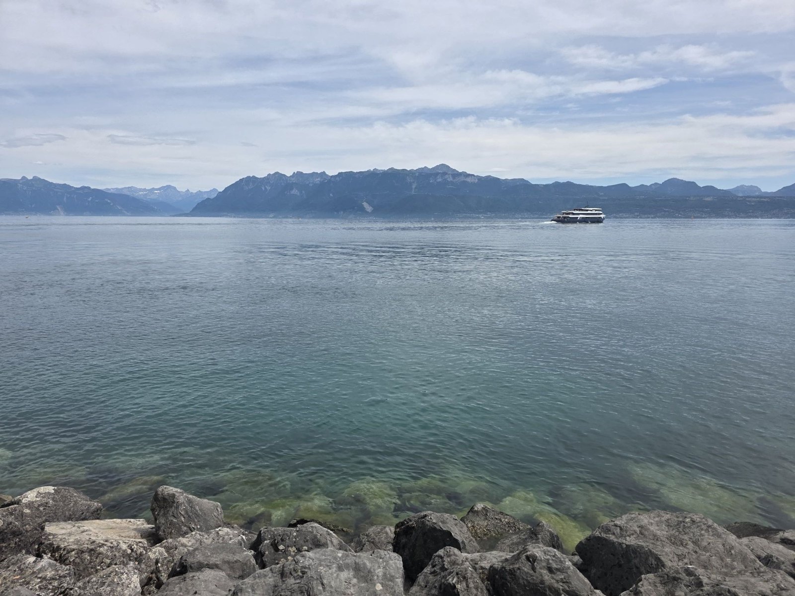 Grey boulders at the water’s edge, clear green water over stones, the lake going blue-grey towards a white passenger boat leaving a wake, and mountains along the far shore under a grey-white sky.