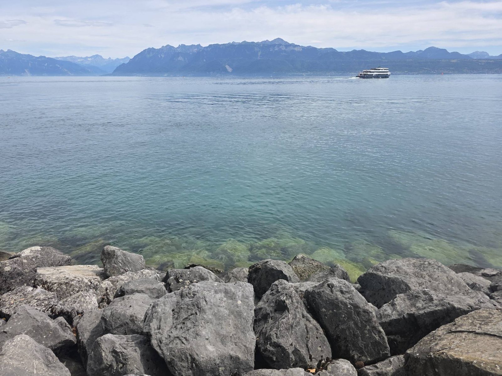 Lake Geneva from a shore of large grey boulders, the water clear green over stones near the rocks and blue further out, a white passenger boat crossing on the right, and a long wall of mountains along the far shore under a pale, hazy sky.