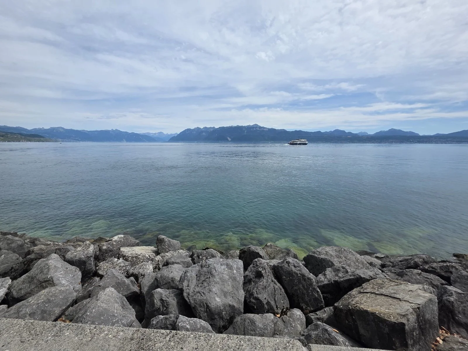 A rocky shore of grey boulders in the foreground, clear green shallows, a wide stretch of blue lake, a white boat in the distance, and the Alps along the whole horizon under a sky of thin cloud.