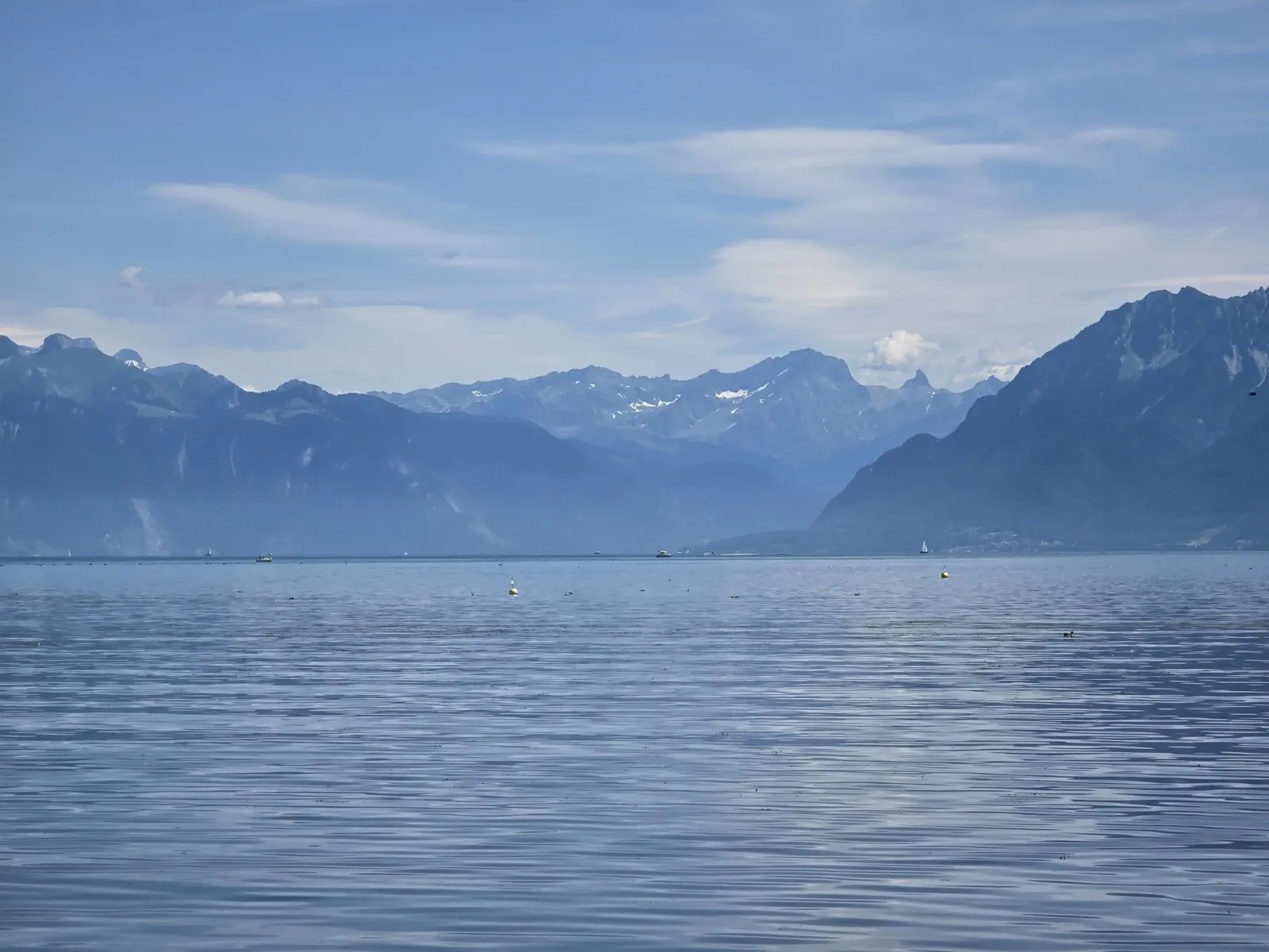 Blue lake water under a blue sky, a yellow buoy and a white one floating near, a few water birds, small boats far off, and across the lake a range of mountains with snow still lying on the highest peaks.