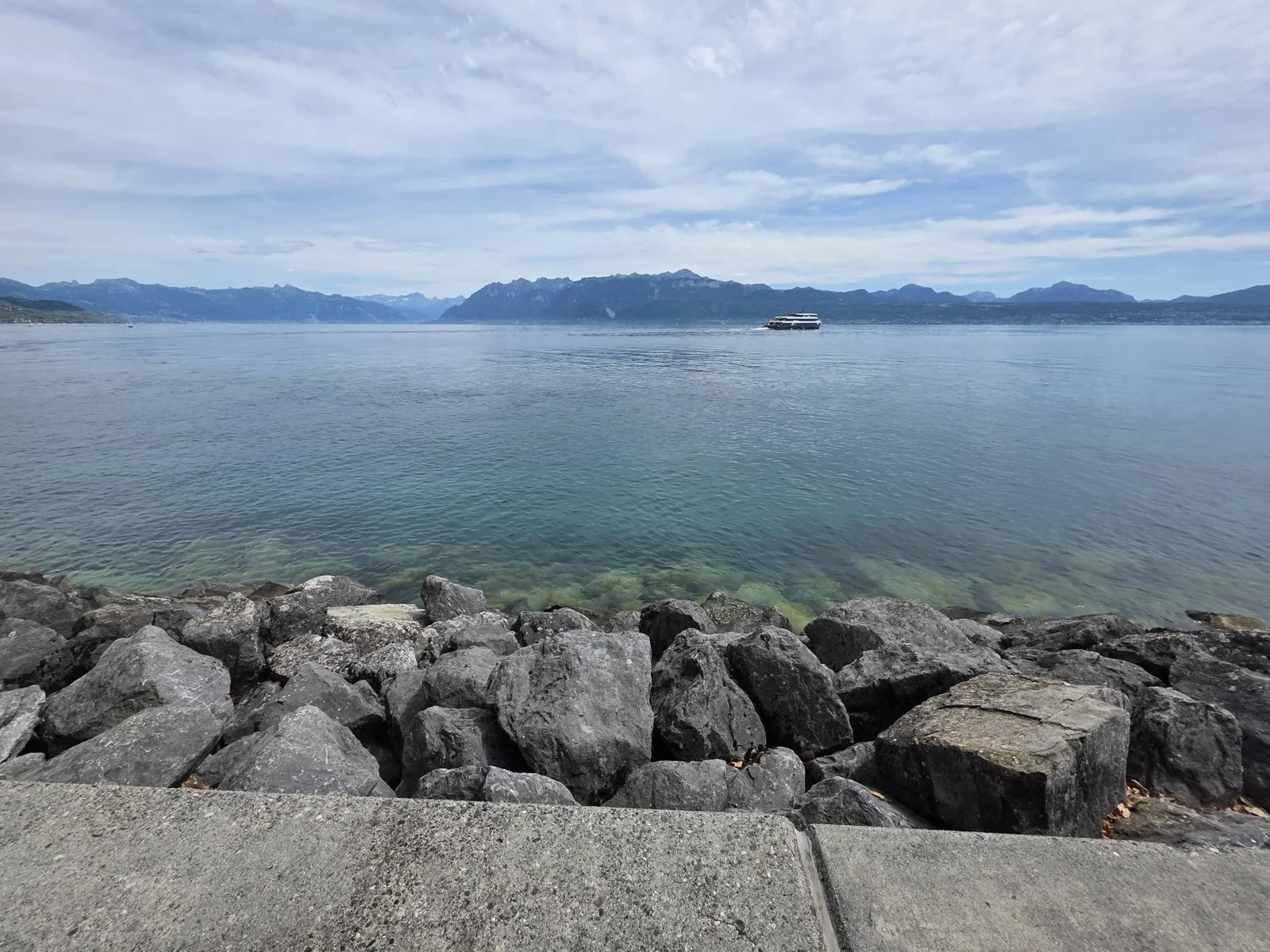 A concrete step and a bank of grey boulders at the lake’s edge, green water in the shallows, a white boat crossing in the distance, and the Alps along the far shore under high cloud.