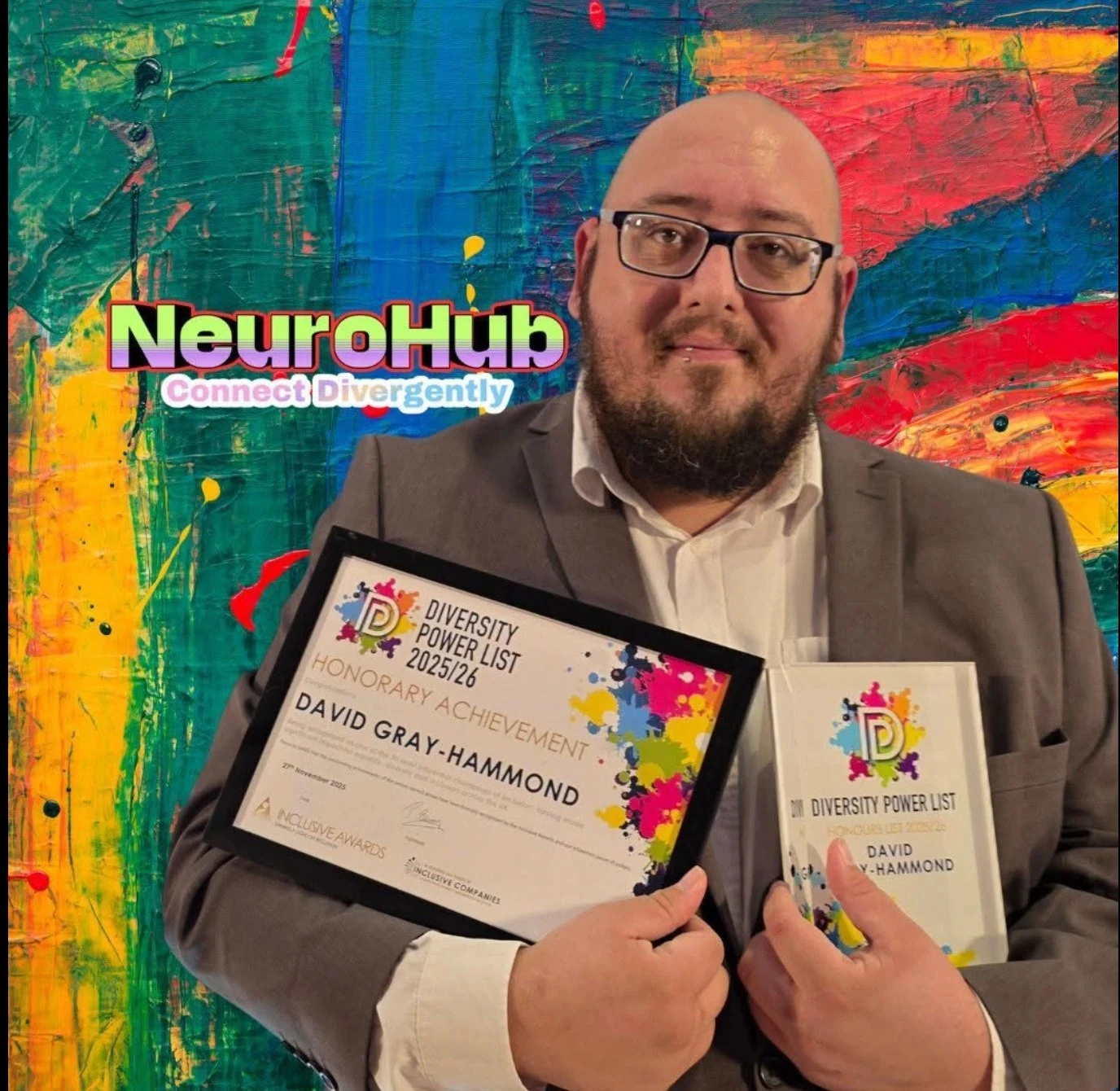 David Gray-Hammond, a bald white man with a dark beard and dark-framed glasses, in a grey jacket over a white shirt, smiling slightly and holding up two awards: a framed certificate reading Diversity Power List 2025/26, Honorary Achievement, David Gray-Hammond, from Inclusive Awards, and a clear block reading Diversity Power List Honours List 2025/26, David Gray-Hammond. Behind him a wall painted in thick strokes of green, blue, red, orange and yellow, with the NeuroHub logo and the words Connect Divergently across it.