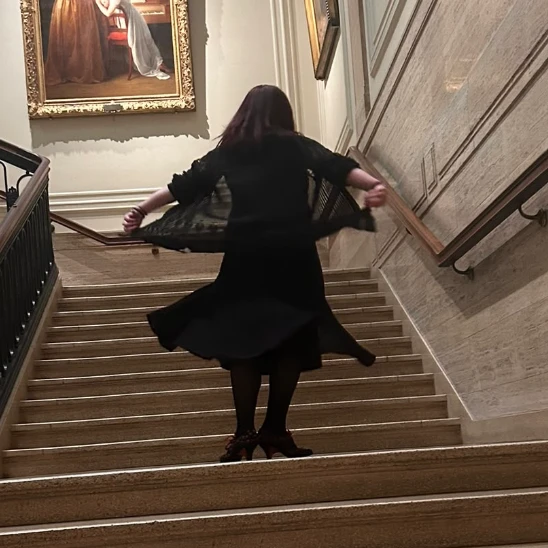 A white woman with reddish brown hair in a flowing black dress ascends a grand staircase.
