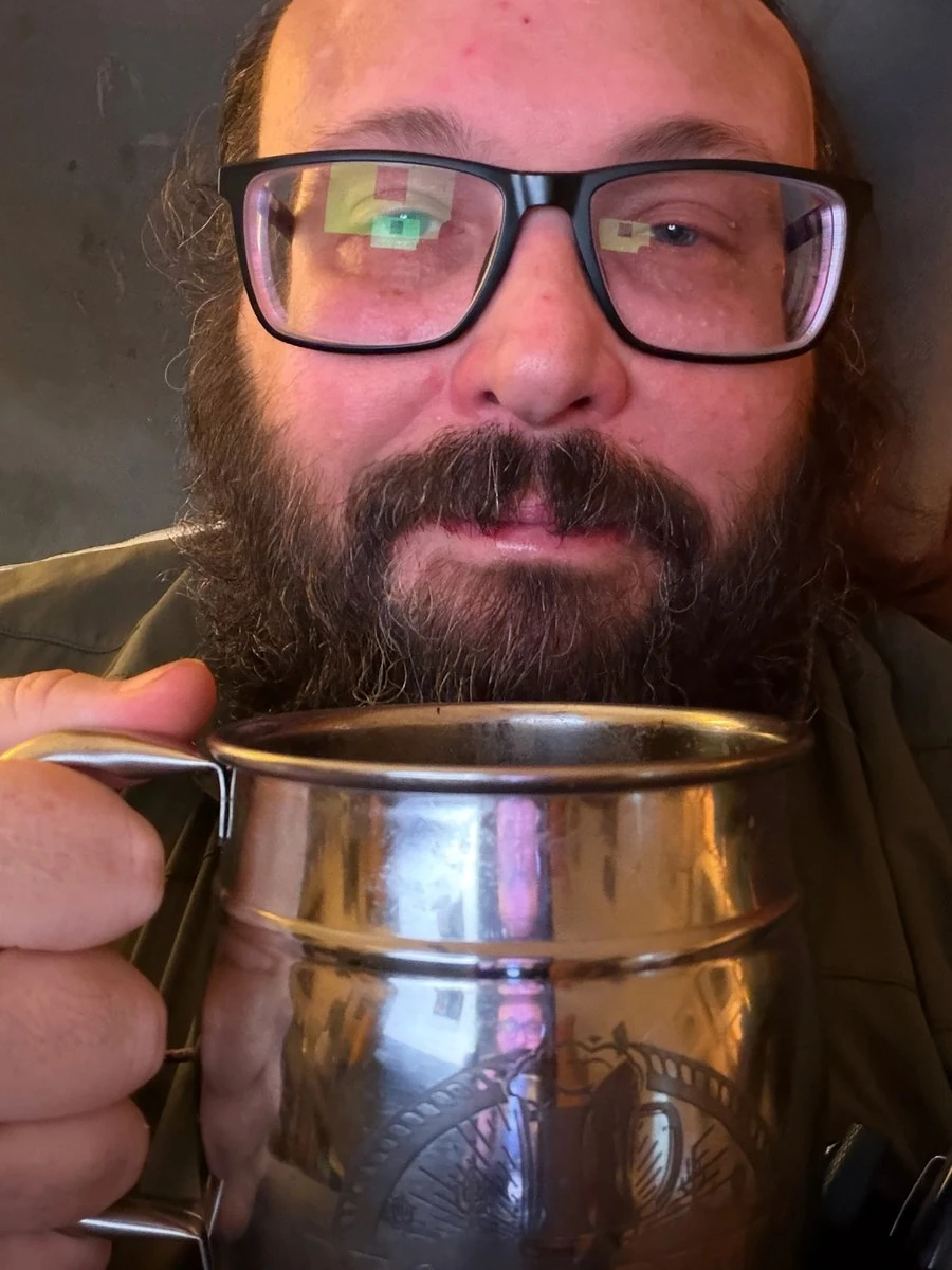 A middle-aged white man with a brown beard and black-framed eyeglasses holds a silver tankard in front of his chin.