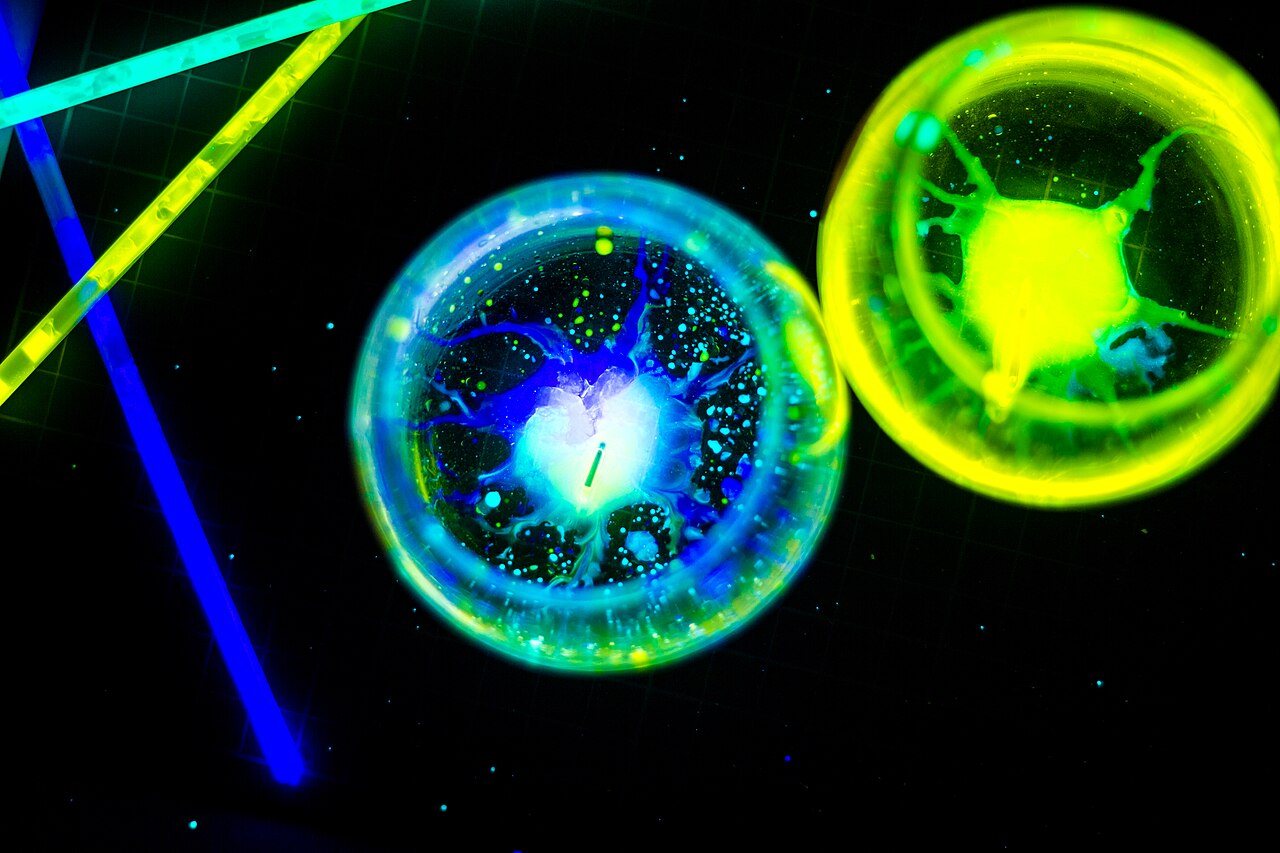 Two round glasses seen from above, glowing blue and yellow-green where glow sticks have leaked into them, with lit glow sticks lying around them in the dark.