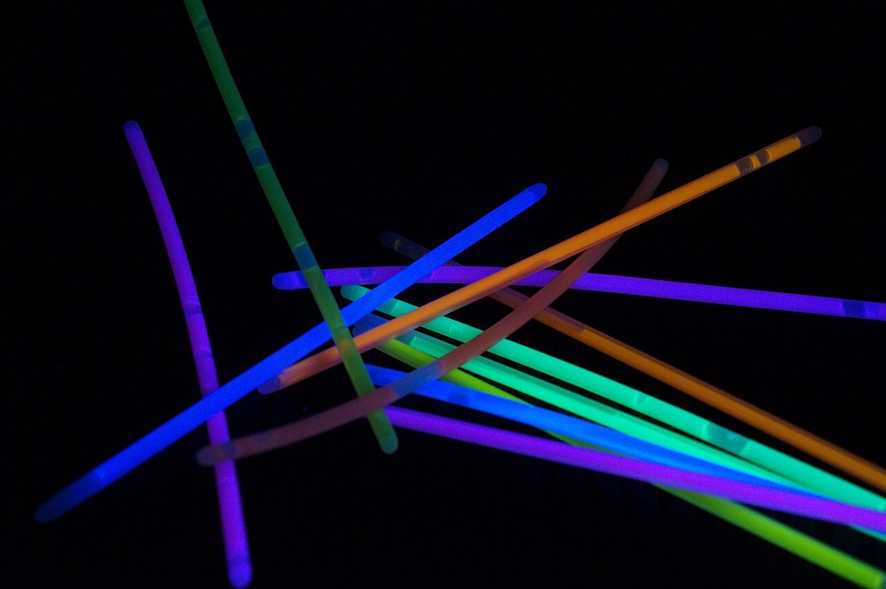 A pile of glow sticks that have not been snapped yet, glowing purple, blue, green and orange under UV light.