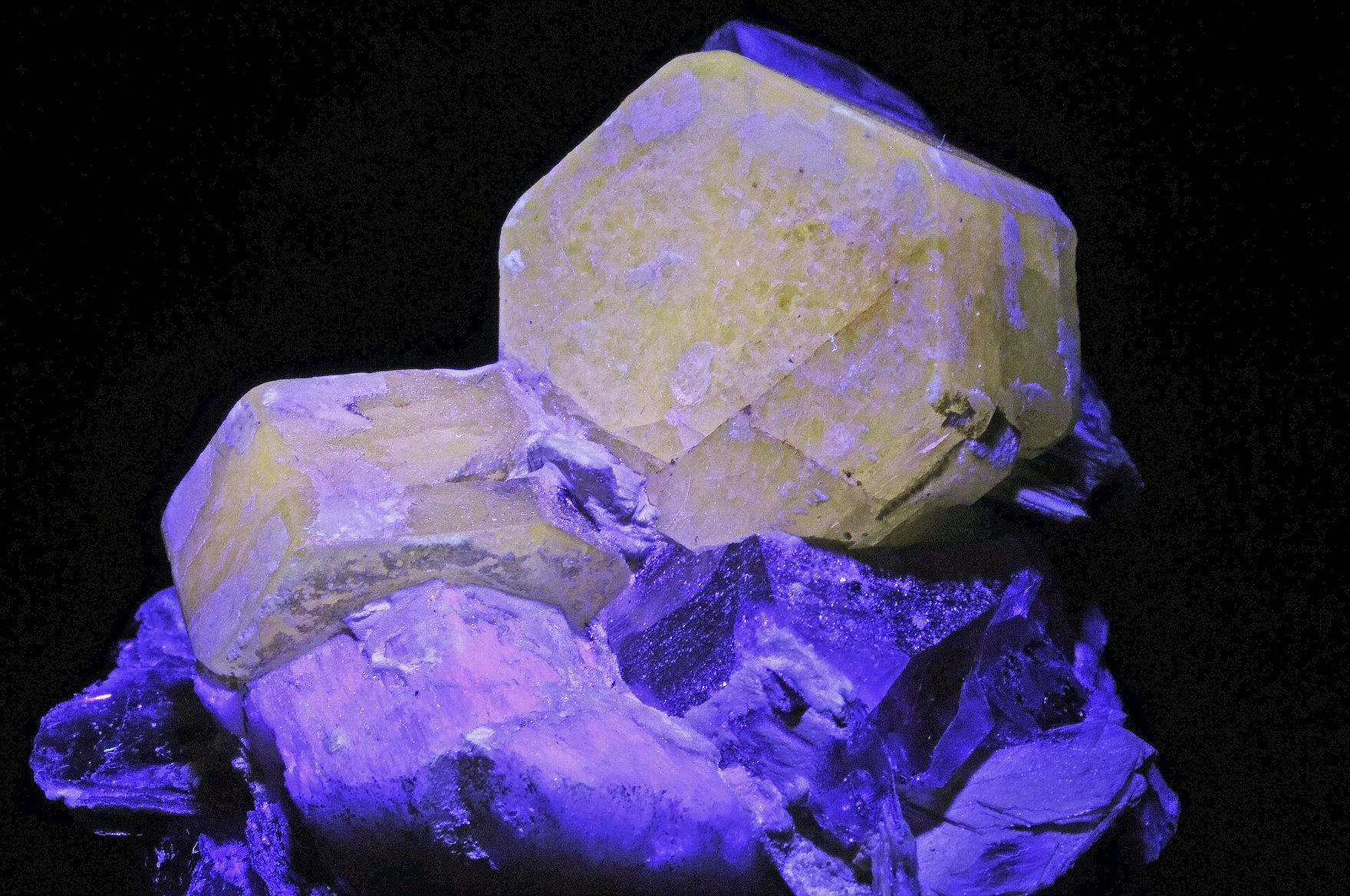 Mineral crystals under UV light, the big ones glowing pale yellow and the rock around them violet.