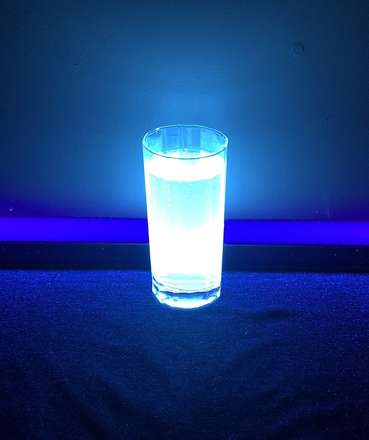 A tall glass of tonic water glowing bright blue-white under a UV tube on a dark cloth.