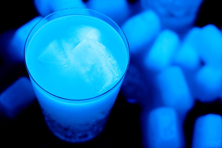 A glass of tonic water with ice cubes made of tonic water, all glowing bright blue under a black light.